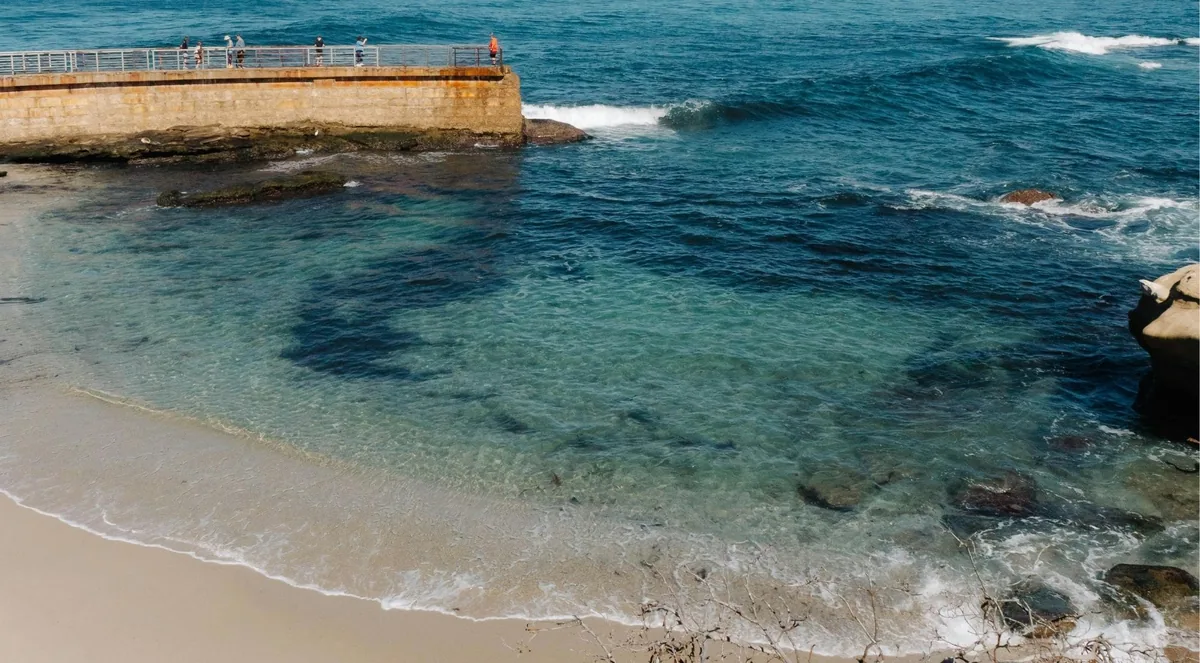 People walking along a curved concrete seawall sheltering a small beach with clear shallows