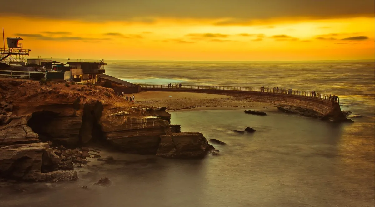 The curved concrete seawall at Children's Pool at sunset, people walking along the rail, a lifeguard tower on the bluff behind