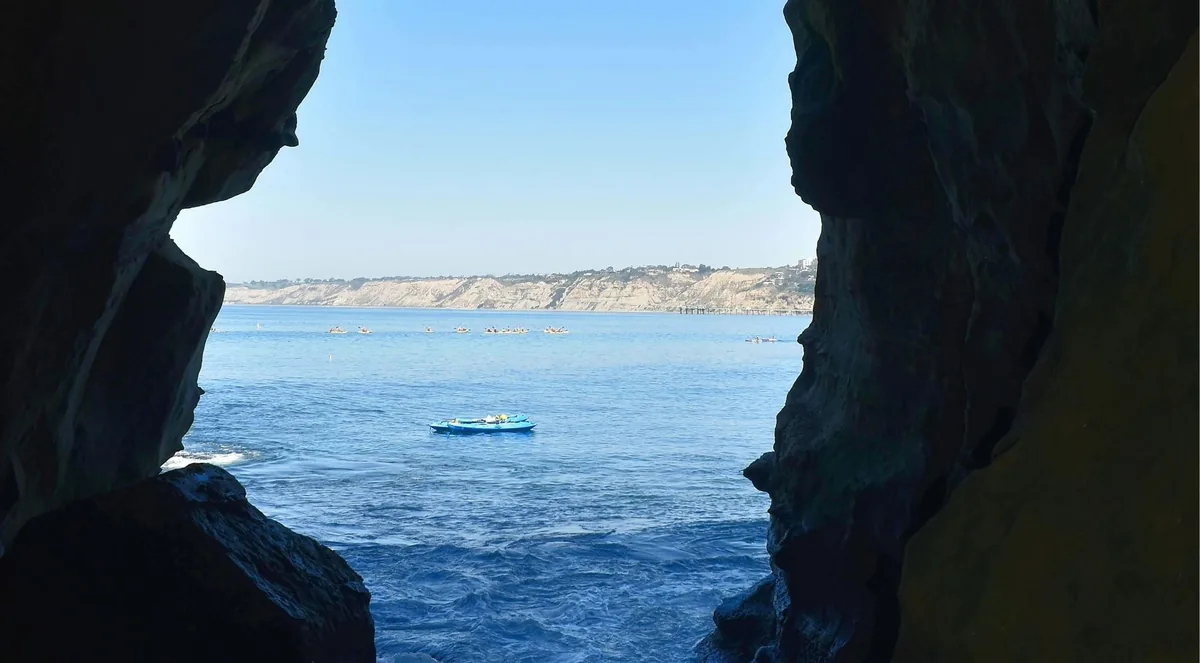 View from inside a dark sea cave out to blue water with a group of kayakers paddling past
